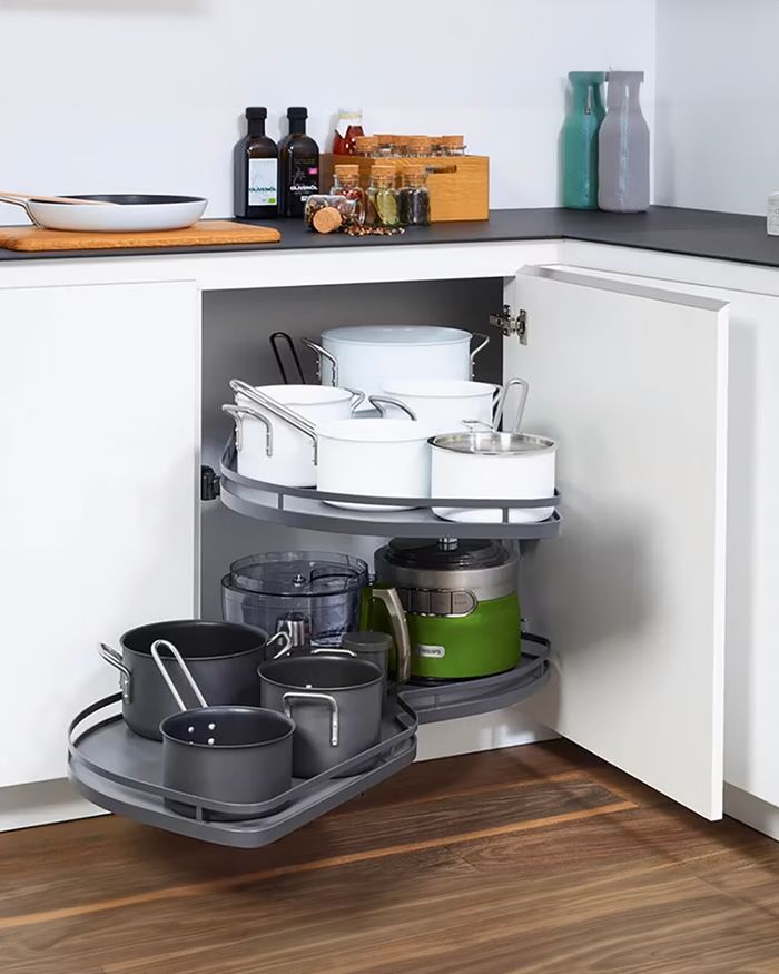 The image features a kitchen corner cabinet by Mebel Arts, equipped with the revolutionary LeMans storage mechanism by Kessebohmer. The mechanism provides two rotating levels, with the lower one accommodating black pots and green-colored electrical appliances, and the upper one containing white kitchen utensils. The cabinet offers a practical solution for organized storage, facilitating access and use while maintaining the style and contemporary aesthetics that characterize Mebel Arts.