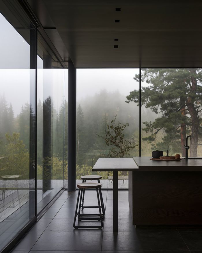 Cuisine moderne avec îlot, tabourets en bois et grandes fenêtres donnant sur une forêt verdoyante.